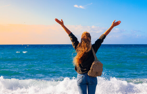 Happy woman with arms raised standing on a pebble beach enjoying the Mediterranean sea at sunset. Freedom, success and travel lifestyle concept. Authentic emotions and coastal nature view.