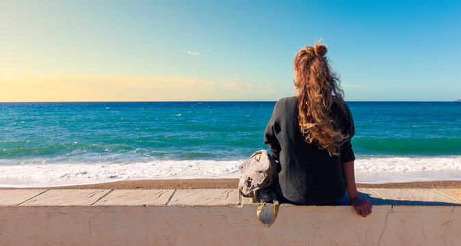 Lonely woman sitting on a wall looking at the Mediterranean sea at sunset. Contemplation, peace and mindfulness travel lifestyle concept. Authentic girl with curly hair on a beach background.