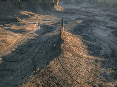 Aerial view of a sharp rock spire rising from the textured badlands and eroded desert terrain under warm golden hour light in Hanksville, Utah, United States.