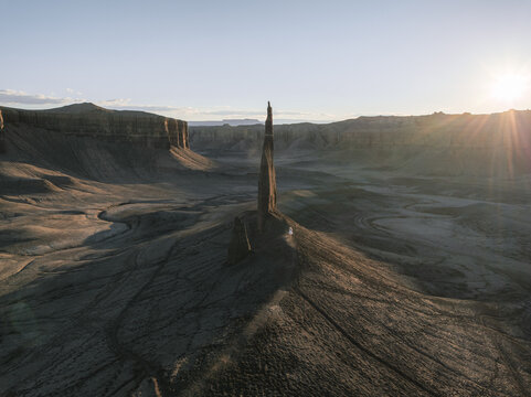 Aerial view of a lone rock spire rising from the desert floor with a person in a white dress standing at its base during sunset in Hanksville, Utah, United States.