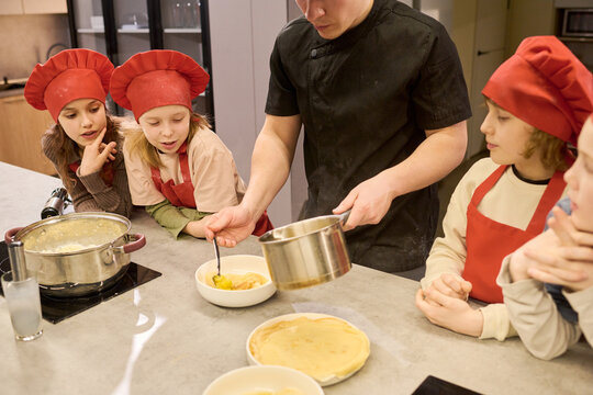 Young adult male chef serving food to children during cooking class, teaching meal preparation skills in kitchen. Useful for culinary education, teamwork, or kids workshop promotion