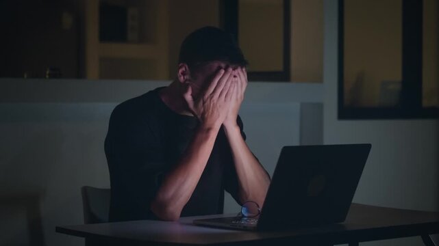 Overworked man using laptop at night takes off glasses and hides face in stress. Overwork concept reflects long hours work pressure fatigue and unhealthy work life balance