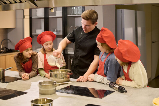 Caucasian male chef teaching children cooking class, pouring ingredient into bowl while kids watching process in training kitchen. Suitable for culinary education, parenting, teamwork marketing