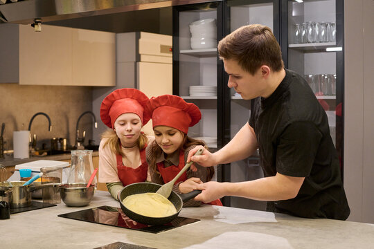 Young man teaching two girls cooking batter in kitchen, supporting learning and practical life skills. Useful for parenting, culinary class, childcare, or education marketing