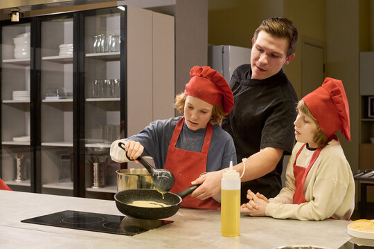 Caucasian male chef guiding kids cooking pancakes in training kitchen, teaching food preparation skills and teamwork. Ideal for culinary class, family cooking program, youth education marketing