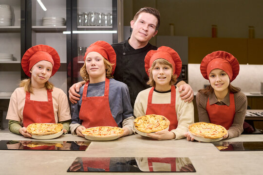 Portrait of Caucasian male chef standing with school age kids holding baked pizzas during cooking class. Useful for culinary education, teamwork, family activity, food workshop promotion
