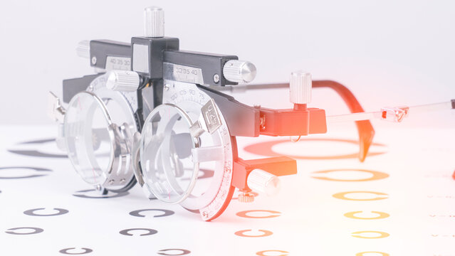 Close-up shows an optometric phoropter with dual lenses. Metallic frame features red and black accents. White background displays circular eye chart symbols. Various markers line the surface