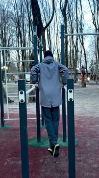 Person seen from behind performing exercises on parallel bars in a park workout area. Surrounded by bare trees, they wear winter clothing, indicating a cold weather workout session