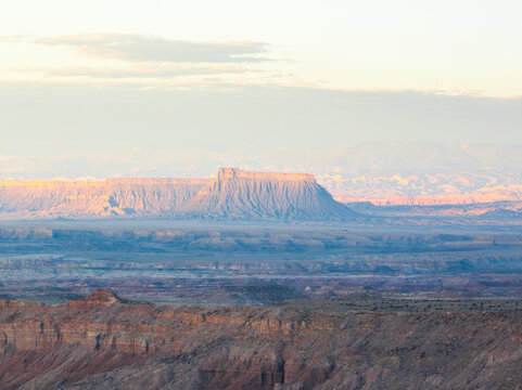 Aerial view of the vast desert landscape featuring prominent mesas and rugged rock formations under a soft golden sky near Goblin Valley State Park in Green River, Utah, United States.
