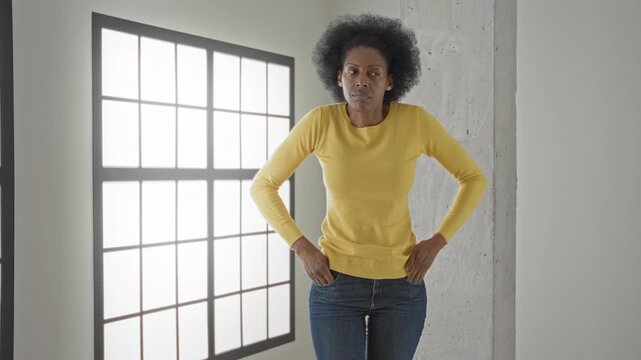 Woman in yellow sweater pulling out empty jeans pockets and shrugging beside large grid window in studio; financial worry.