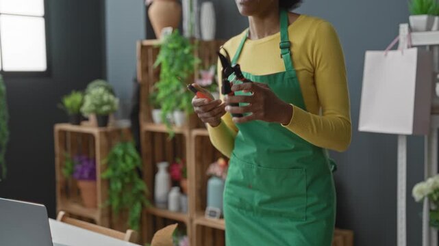 Woman florist in green apron holding pruning shears and trimming stems at a flower studio counter with wooden crates and vases; calm focus.