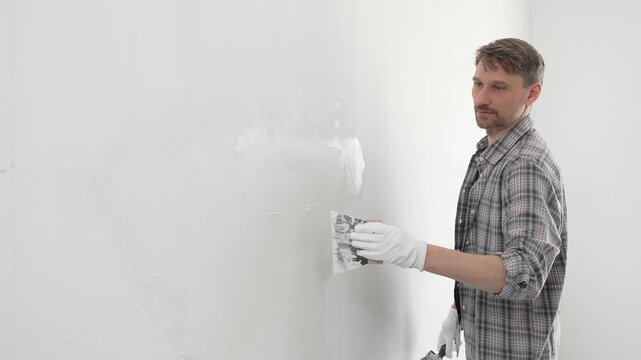 Construction worker wearing gloves and using a putty knife to plaster a white wall, performing home renovation and improvement tasks with precision