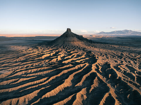 Aerial view of the prominent Factory Butte rising above intricate eroded badland ridges under a clear sky during golden hour in Wayne County, Utah, United States.
