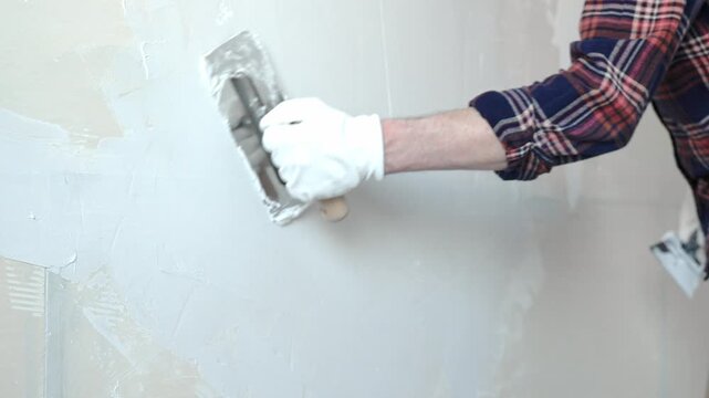 Unknown male construction worker wearing protective gloves smoothing plaster on interior wall during home renovation, applying material with professional trowel technique, close-up