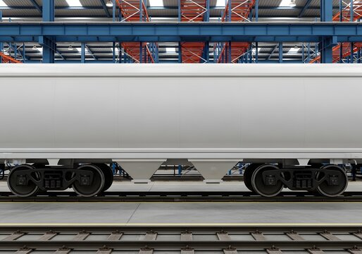 Side view of a white rail hopper wagon inside an industrial facility with blue steel frame structures, orange storage racks, and concrete flooring with integrated rail tracks.