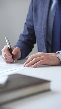Close up of businessman's hands signing a contract with elegant pen, wearing a blue suit and a wristwatch, sitting at a white desk. Business people concept
