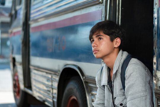 A teenage boy wearing a gray hoodie sits beside a local bus station, carrying a backpack. He looks ahead with a thoughtful and slightly serious expression.