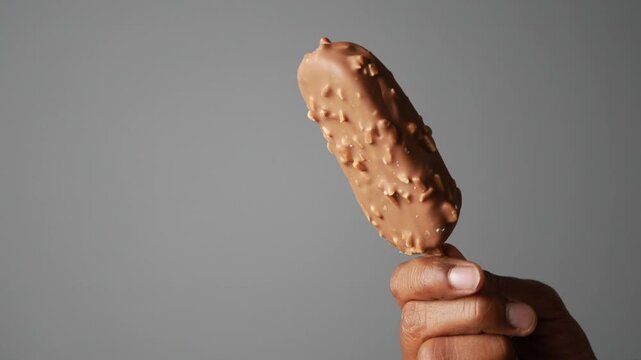 Person holding almond chocolate ice cream bar on stick against gray background slow motion.