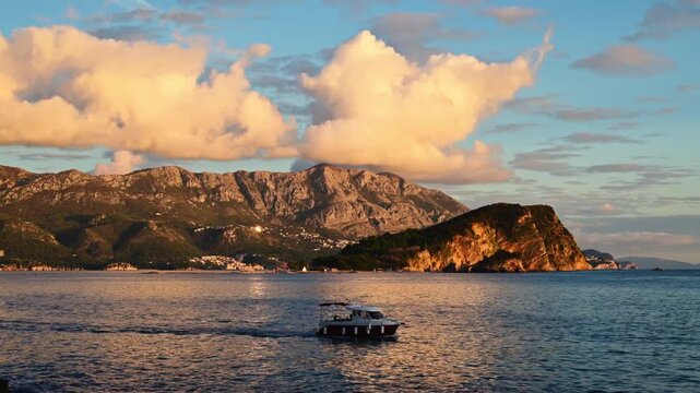 Small motorboat glides over tranquil water with majestic mountains and clouds at sunset.