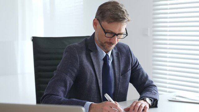 Adult businessman wearing blue suit and tail is concentrating while writing on a document at his desk in a bright, modern office, demonstrating professionalism and efficiency. Business people concept
