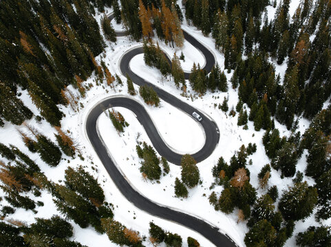 Aerial view of Strada a serpente Passo Giau winding through a snow-covered pine forest with a car on the road in Cortina d'Ampezzo, Veneto, Italy.