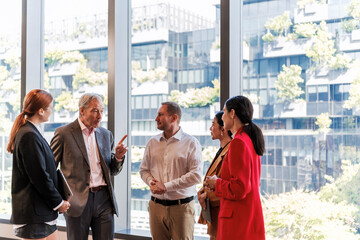 Fototapeta premium A diverse group of professionals holds a business discussion by large office windows overlooking a modern, greenery-covered building.
