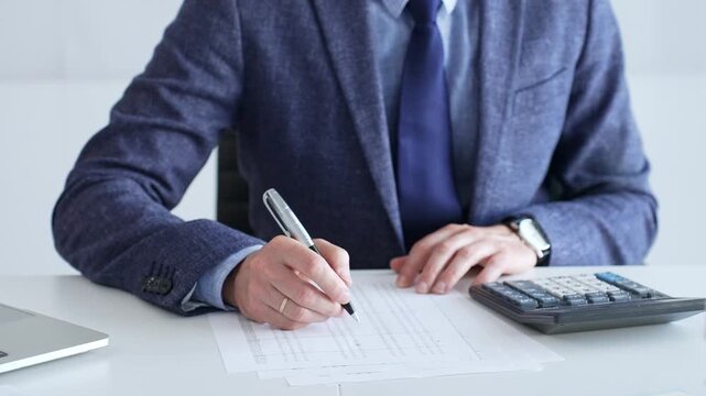 Businessman wearing suit and tie is using calculator and taking notes while working at desk in office, with laptop and stack of folders nearby, close up. Audit and taxes theme in business