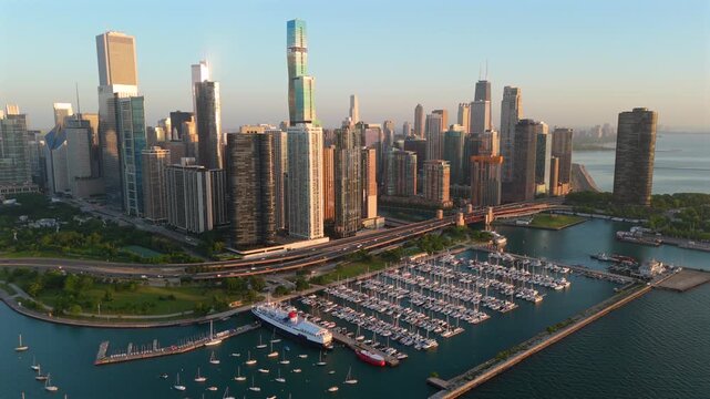 Stunning aerial drone footage of Chicago downtown skyline at golden hour featuring marina with sailboats and yachts along Lake Michigan waterfront, elevated highway traffic, and gleaming skyscrapers.