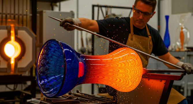 Professional glassblower shaping glowing molten glass in a workshop. Artisan uses a blowpipe to manipulate vibrant orange and blue glass near a furnace. Traditional craftsmanship, creative artistry.