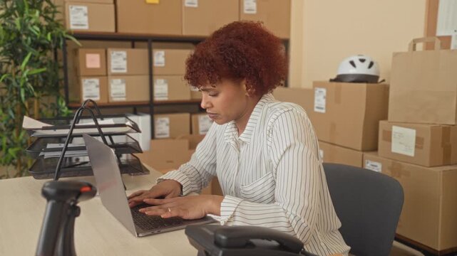 Woman at small business packing desk types on laptop and holds lower back amid stacked cardboard boxes in a building; strain.