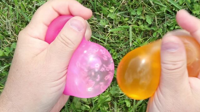 Hands squeeze and press two transparent squishy balls pink and orange on green grass outdoors in daylight ASMR antistress satisfying sensory toy video