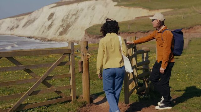 Two hikers pause by a rustic gate overlooking the sea. Sunlit cliffs rise behind them, calm and wild. They gaze at the waves, quiet and content. Perfect for travel or nature-themed projects
