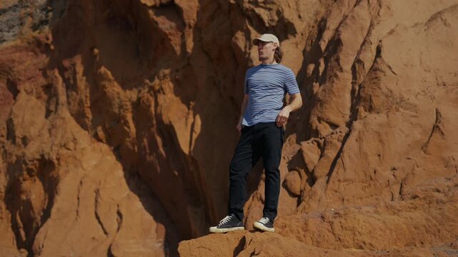 Man stands on rocky ledge, shielding eyes from sun. He shifts stance, surveying expansive red canyon walls. Bright light enhances rugged textures and earthy tones. Ideal for adventure, outdoor