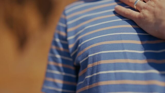 Hand rests gently on chest, conveying calm or introspection. Ring glints subtly, adding personal touch to gesture. Striped shirt shows comfort, warmth, and quiet resolve. Ideal for scenes of peace