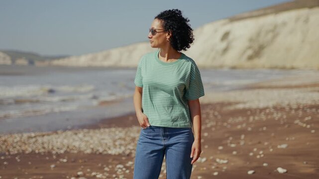 Woman stands relaxed on sunny beach. She gazes at ocean with quiet enjoyment. Waves crash gently against pebbled shore. Cliffs rise in soft, sunlit backdrop. Perfect for travel, lifestyle