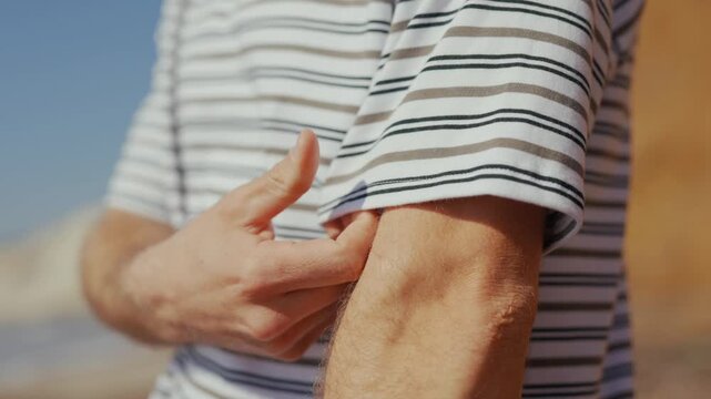 Man adjusts striped shirt sleeve. Hand grips fabric near elbow. Sunlight highlights skin texture. Scene feels relaxed and casual. Perfect for beach or summer themes. No animals or plants visible
