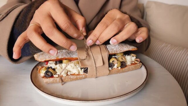 Woman hands untie string on rustic baguette sandwich with cheese and olives.
