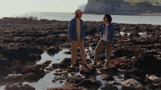 Sunlight glints off tidal pools between rocks. Two people stand on wet, mossy stones near sea. They chat softly, enjoying the coastal breeze. Perfect for romantic or adventurous travel scenes