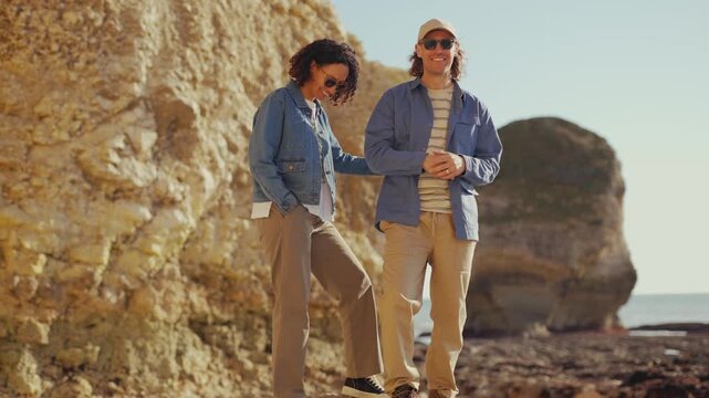 Two friends stand near a rocky coast. They wear casual, matching jackets. One adjusts sunglasses, the other smiles faintly. Sunlight warms the golden cliffs behind them