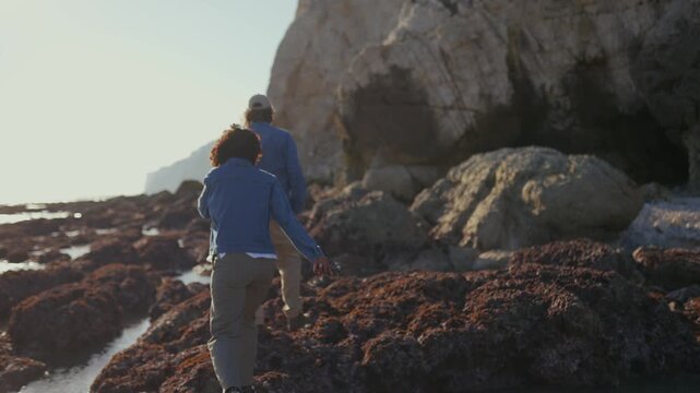 Two people explore rocky, seaweed-covered coastline. Sunlight casts long shadows across wet stones and cliffs. They walk slowly, curious about the tide pools and caves
