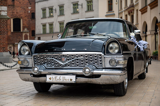 A Gaz-13 Chaika is parked in Krakow, Poland with wedding decorations and old buildings visible