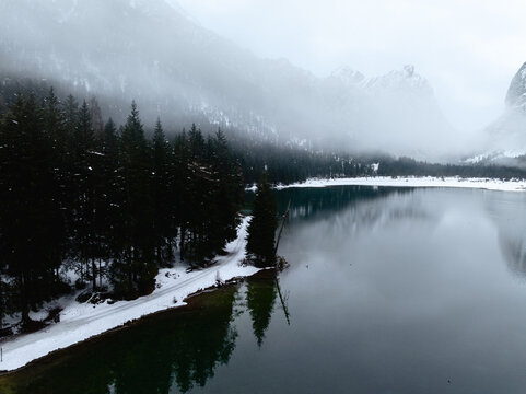 Aerial view of Toblacher See with snow-covered shores, dense pine forest, and misty mountains in Toblach, Trentino-South Tyrol, Italy.