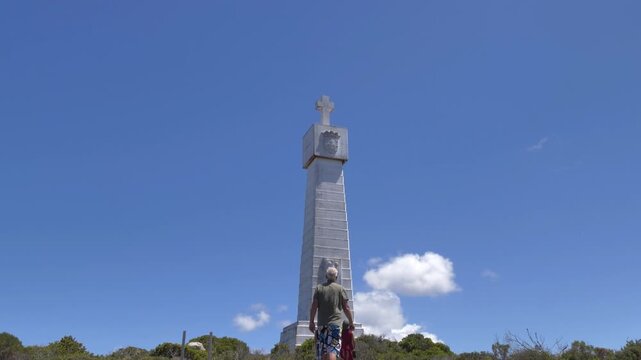 A scenic view of Dias Cross, a prominent monument, at Cape Point, Cape Point Nature Reserve, with a few people approaching it on a sunny day.