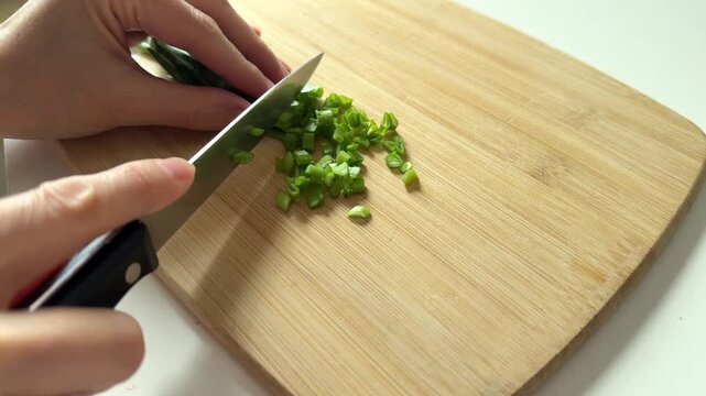 Person cutting fresh green onion on wooden cutting board with knife. Food preparation, cooking, fresh ingredients