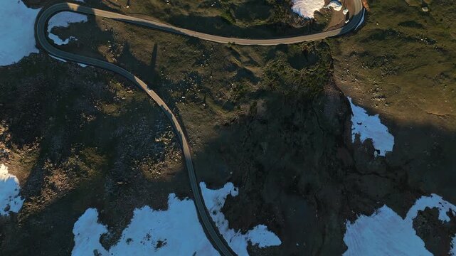 Aerial drone footage captures winding serpentine mountain road cutting through rugged alpine terrain with patches of melting snow scattered across dark green and brown landscape during warm golden.