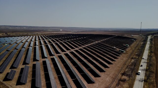 Aerial view of solar panel farm showing rows of solar panels aligned in a field with vehicles parked along a road under a clear blue sky in a rural area