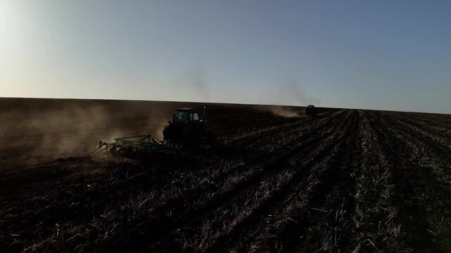 Two tractors plowing a large field in a rural area, creating dust clouds as they work in parallel rows under a clear blue sky during the day