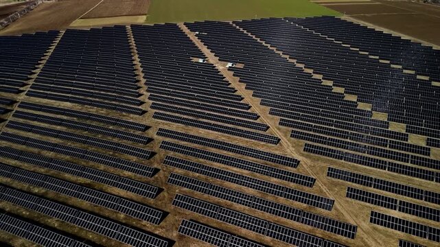Aerial view of expansive solar panel farm with rows of photovoltaic panels arranged in a grid pattern under clear blue sky and surrounding agricultural fields
