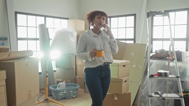 Woman holding yellow mug and smartphone to ear in a building, smiling while unpacking moving boxes and surveying her new home; settling in contentment.