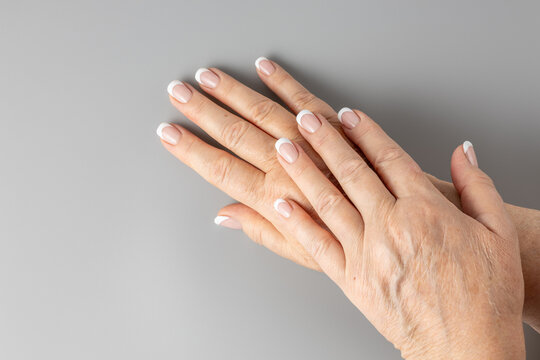 Profesional french nails manicure on aging hands of adult 65 year old woman with age spots, deep wrinkles on dry skin. Skin aging and elderly nail care concept. Female hands on gray background.